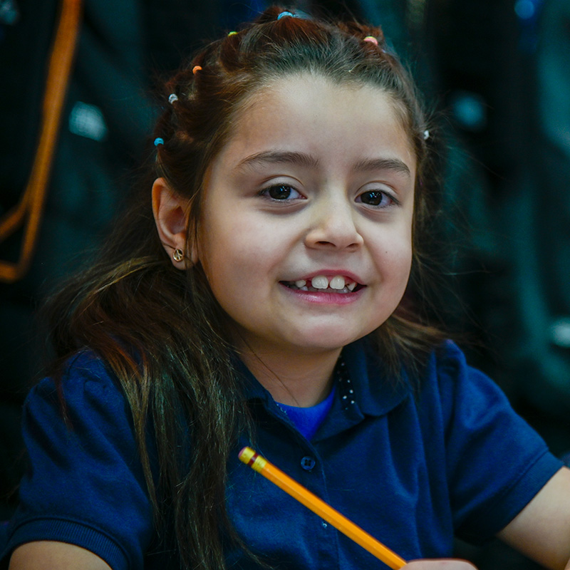 A young girl holding a pencil smiles in her classroom